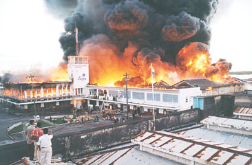 Winston Oudkerk photo shows Muneshwers building in flames. Inset: The elder Muneshwer (center) contemplating his next move as he and family members look at the raging inferno.